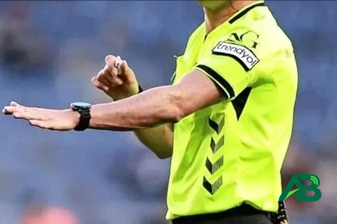 Sarı forma giyen futbol hakemi sahada elinde düdükle işaret veriyor.