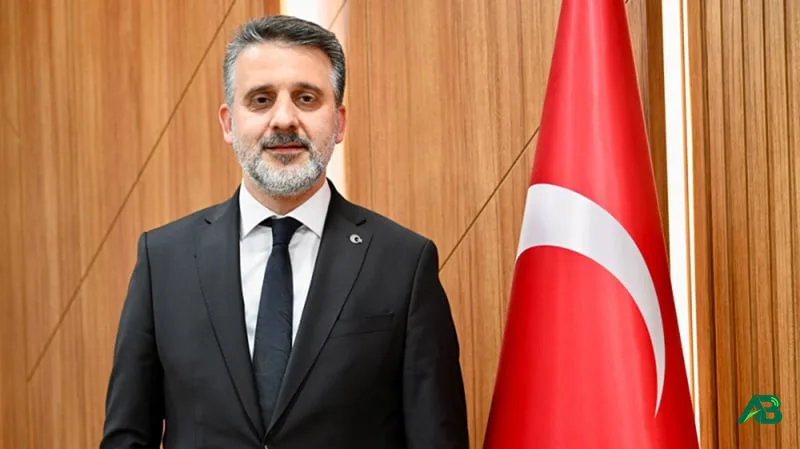 Siyah takım elbiseli ve kravatlı bir erkek, arkasında Türk bayrağı ve ahşap panellerle poz veriyor.