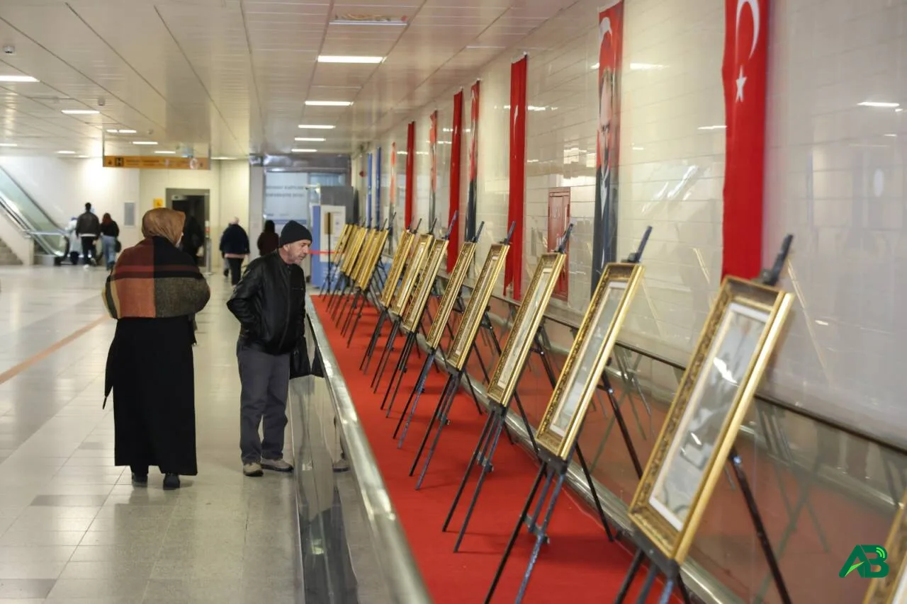 Kapalı bir alanda şövale üzerinde sergilenen tabloları inceleyen insanlar ve duvarda asılı Türk bayrakları.