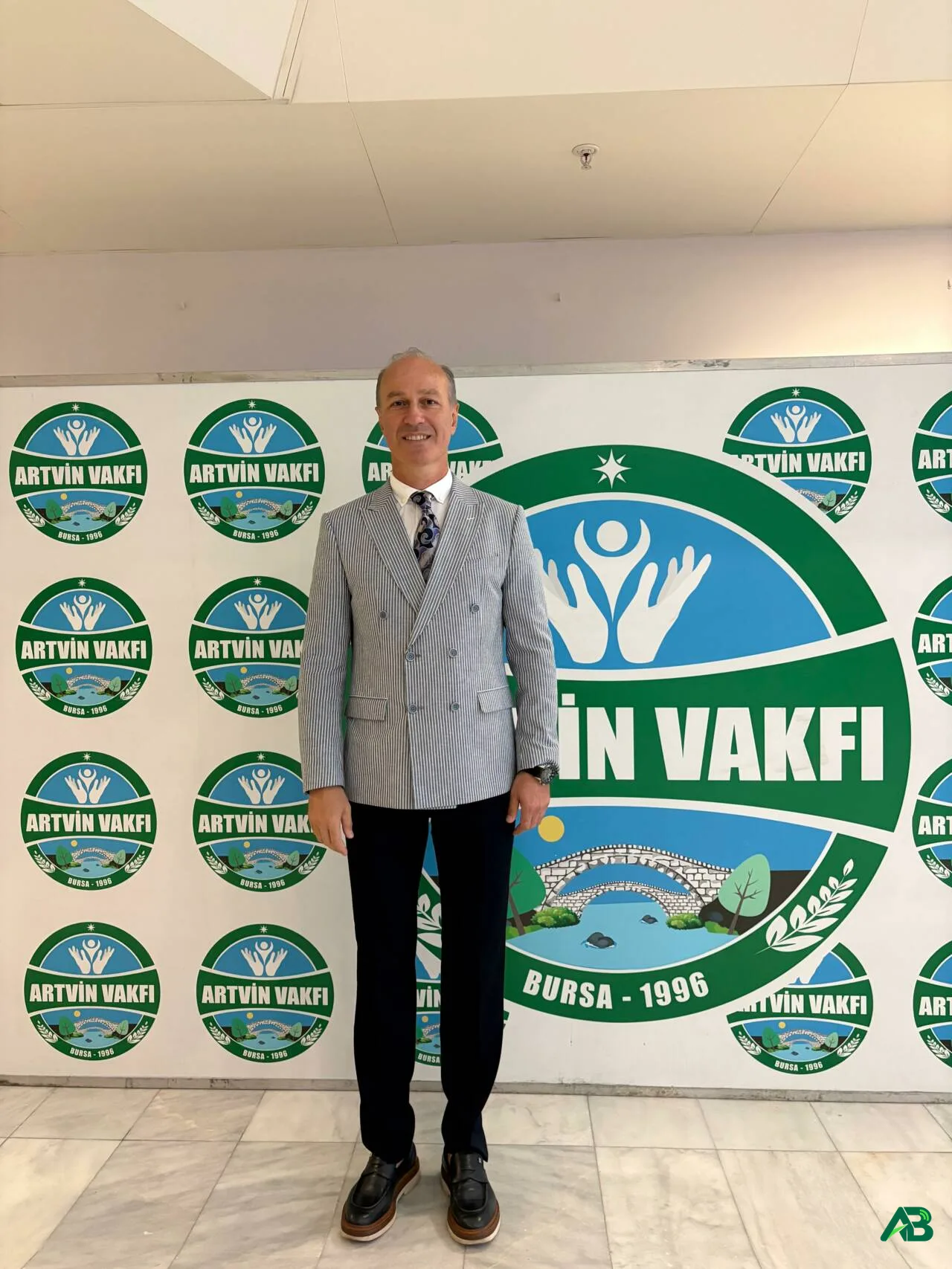 Bursa Artvin Vakfı logolu arka plan önünde duran takım elbiseli bir erkek.